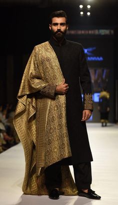 a man is walking down the runway wearing a black and gold outfit with an elaborate shawl