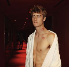 a shirtless young man standing in a hallway wearing a white robe and looking at the camera