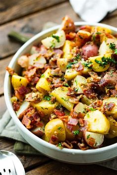 a white bowl filled with potatoes and bacon on top of a wooden table next to utensils