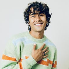 a young man with curly hair wearing a green and orange striped sweater smiles at the camera