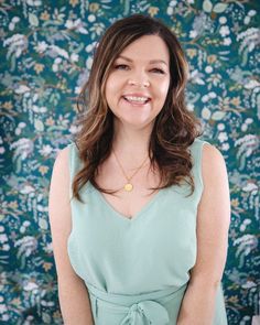 a woman in a green dress smiling at the camera with floral wallpaper behind her