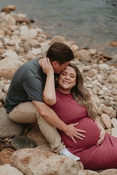 a pregnant couple cuddling on rocks by the water