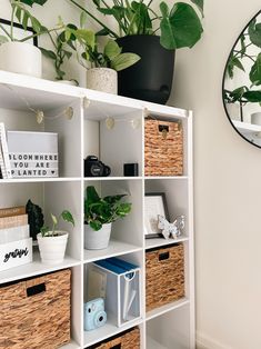 a white bookcase with baskets and plants on the top, next to a mirror