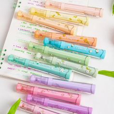 five different colored pens sitting on top of each other next to green leaves and flowers