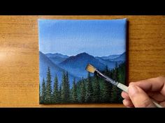 someone is holding a paintbrush and painting a landscape with mountains in the back ground