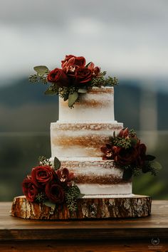 a three tiered cake with red flowers and greenery sits on a wooden table