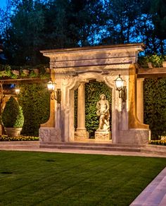 an outdoor area with a statue and lights in the background at night, surrounded by greenery