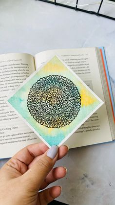 a hand holding an open book on top of a table with watercolor and ink