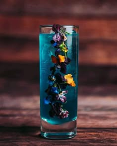 a tall glass filled with blue liquid and colorful flowers on the rim, sitting on a wooden table