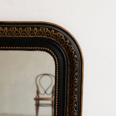 an ornate black and gold framed mirror against a white wall with a chair in the reflection