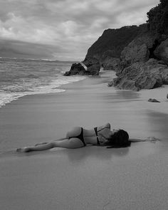 a woman laying on top of a sandy beach next to the ocean with rocks in the background