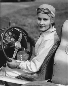 a woman sitting in the driver's seat of an old race car with her hands on the steering wheel