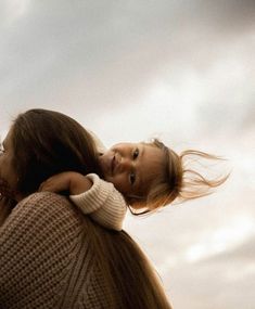 a woman holding a small child in her arms and looking up at the sky with clouds behind her