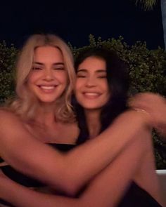 two beautiful women hugging each other in front of a fire pit at night with palm trees behind them