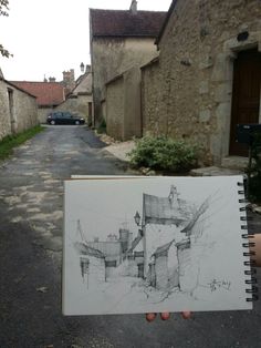 a person holding up a drawing in front of an alley way with cars parked on the side