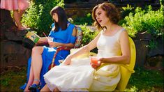 two women in dresses sitting next to each other and one is holding a drink while the other holds a book