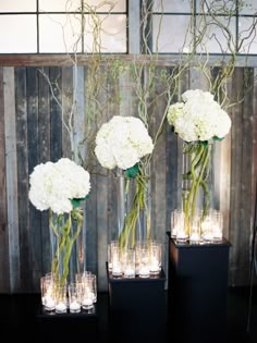 three tall vases filled with white flowers on top of a black table next to candles