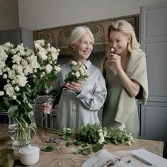 two women standing next to each other with flowers in front of them and an open book on the table