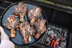 someone is holding up some meat on a plate over an open fire pit with the grill in the background