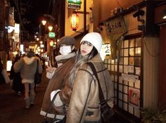 a woman wearing a white hat and coat walking down a street at night with people in the background