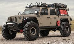 a jeep with large tires is parked on the gravel in front of an open field