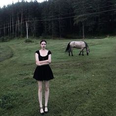 a woman in a black dress standing next to a horse on a lush green field