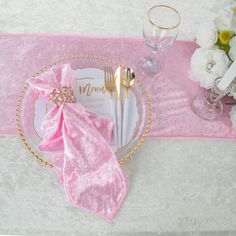 a place setting with pink napkins and goldware on a white table cloth, surrounded by flowers