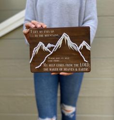 a woman holding up a wooden sign with mountains on the front and back, that says i lift my eyes up to the mountain