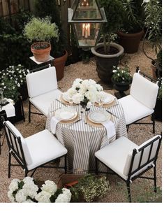 an outdoor dining table set with white flowers and place settings for four in front of potted plants