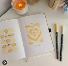 an open book on a table with markers and pens next to it, along with a candle