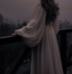 a woman in a long white dress standing on a balcony looking out at the trees