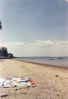 an orange and white blanket is laying on the sand at the beach with boats in the water