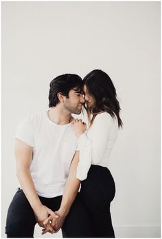 a man and woman sitting next to each other in front of a white wall with their arms around each other