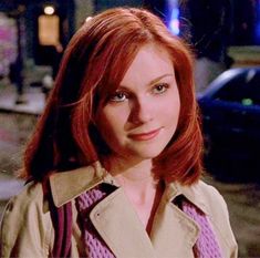 a woman with red hair wearing a trench coat and looking at the camera on a city street