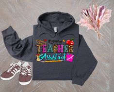 a sweatshirt with the words teacher assistant on it next to a pair of tennis shoes