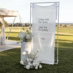 a white sign sitting on top of a lush green field next to a vase filled with flowers