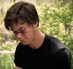 a young man is looking down at his cell phone while standing in the woods with trees and bushes behind him