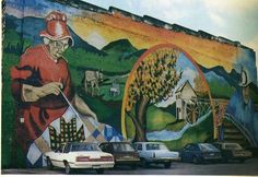 a mural on the side of a building with cars parked in front of it and a man holding a paintbrush