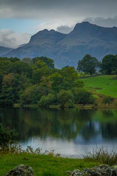 a lake surrounded by lush green trees with mountains in the background on a cloudy day