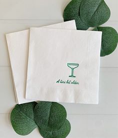 two white napkins with green lettering on them sitting next to some leafy greens