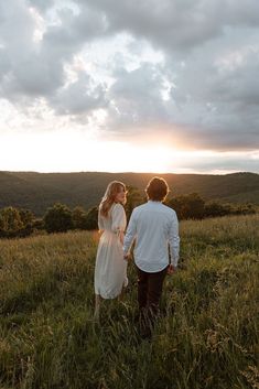 a man and woman holding hands walking through the grass in front of some hills at sunset