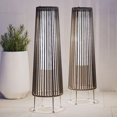 two tall metal vases sitting next to each other on a white tile floor with a plant in the middle