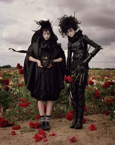 two women dressed in black standing next to each other on a field with red flowers