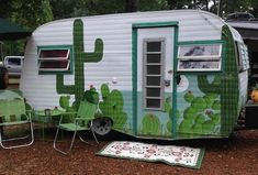 an rv with cactus painted on the side and green lawn chairs next to it,