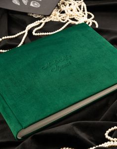 a green book is laying on a black cloth next to pearls and a white beaded necklace