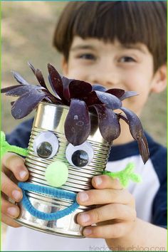 a young boy holding up a tin can with flowers in it and eyes painted on the side