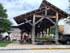 people are standing under a wooden structure with plants on it and cars parked in the background