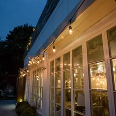 the outside of a restaurant at night with string lights on the windows and patio furniture