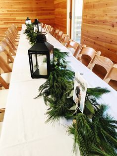 a long table is set up with two lanterns and greenery on it, surrounded by chairs