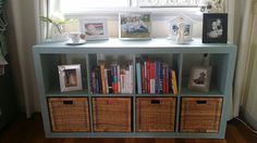 a shelf with books and pictures on it in front of a window filled with wicker baskets